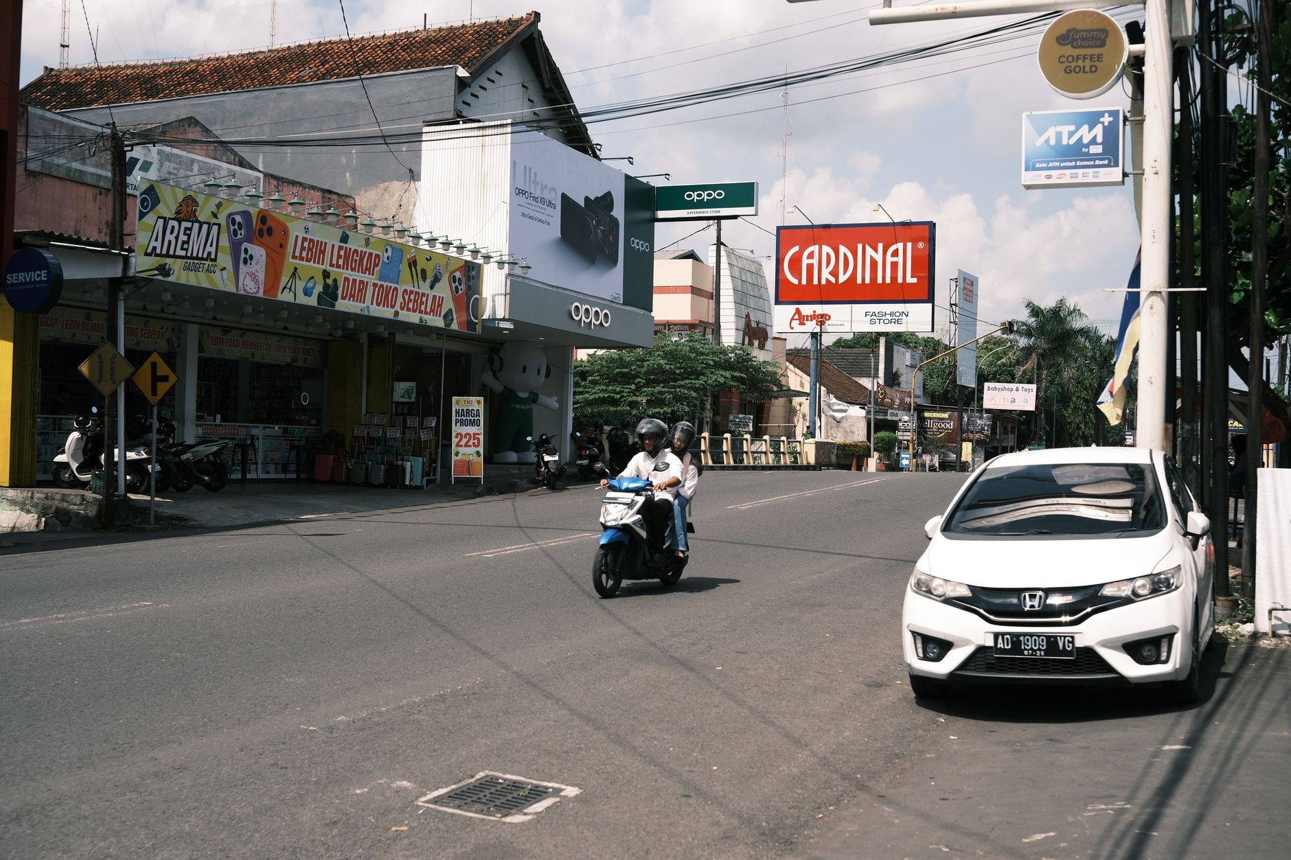 Busy Southeast Asian street with motorcycles and a parked car near shops and billboards.