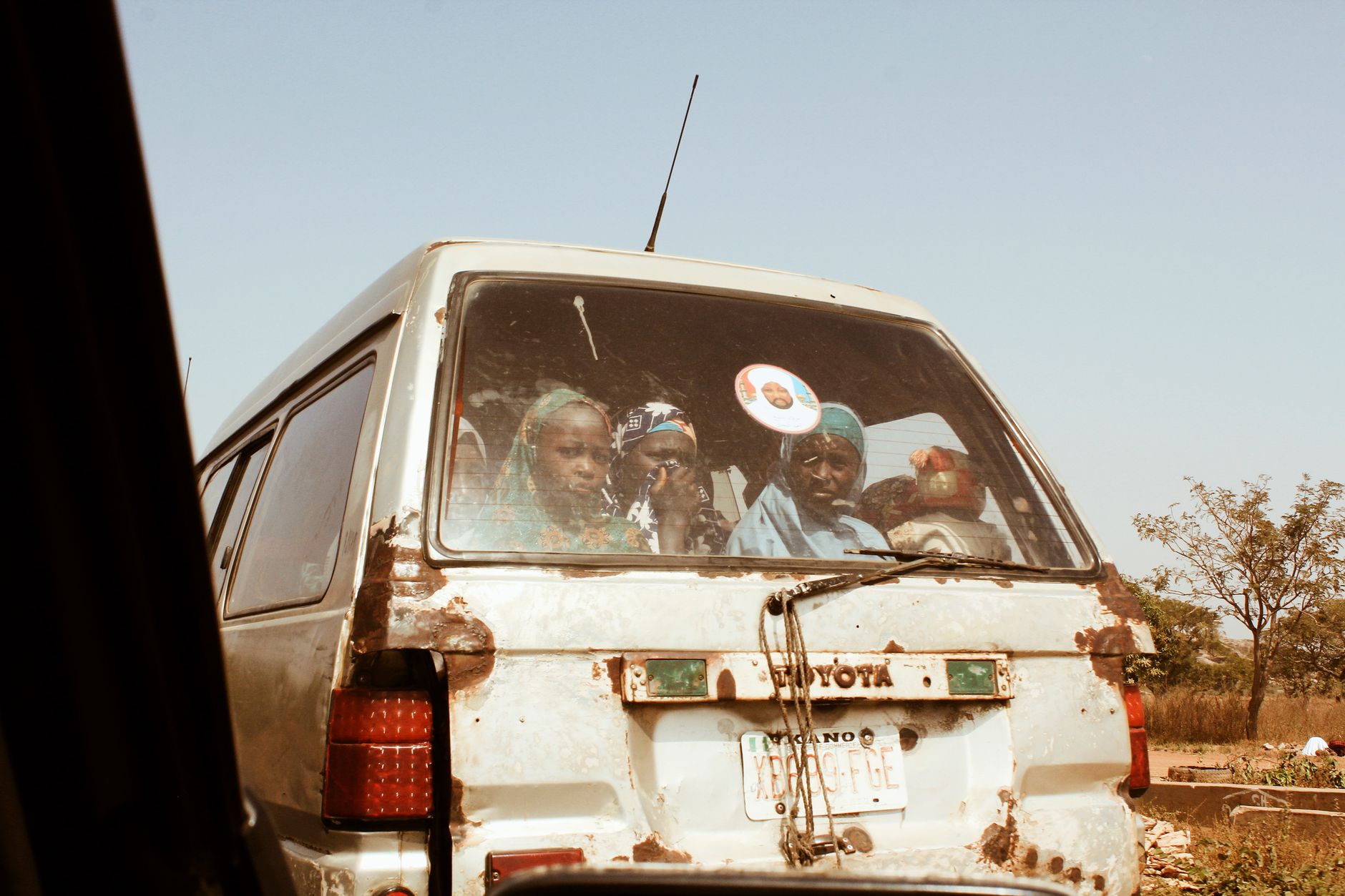 A weathered vintage Toyota van carrying multiple passengers travels through a rural landscape under a clear sky.