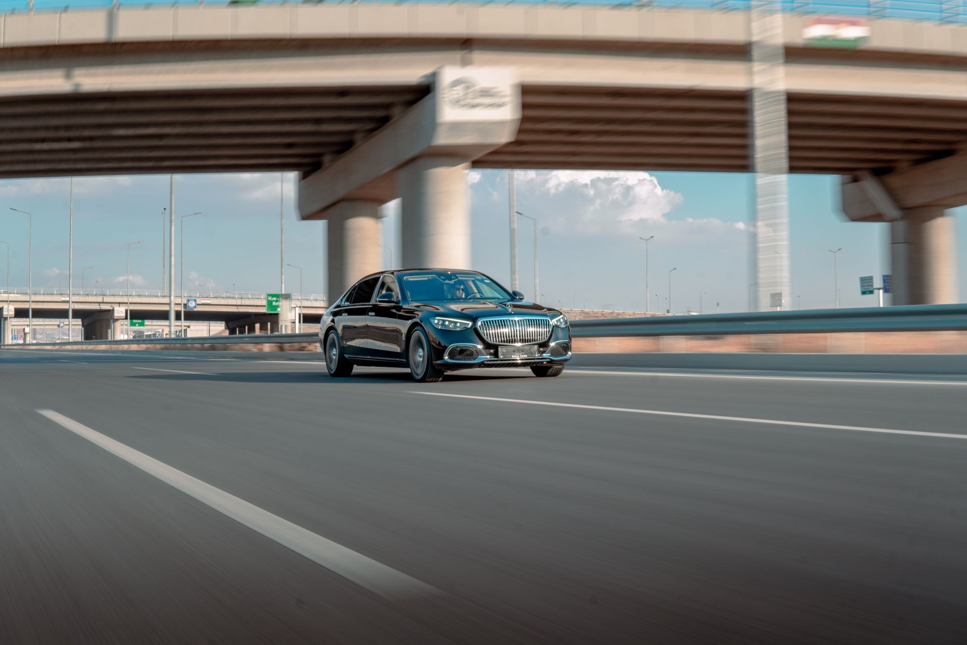 A sleek luxury car traveling fast on an open highway beneath an overpass. Clear blue sky day.