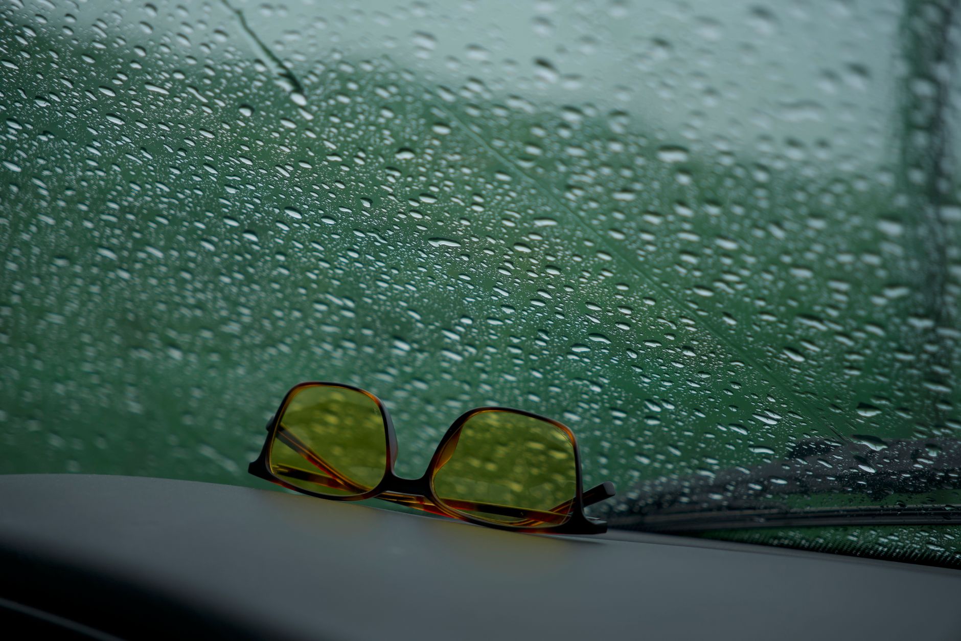 Yellow tinted sunglasses resting on a car dashboard with raindrops on the windshield, creating a moody atmosphere.