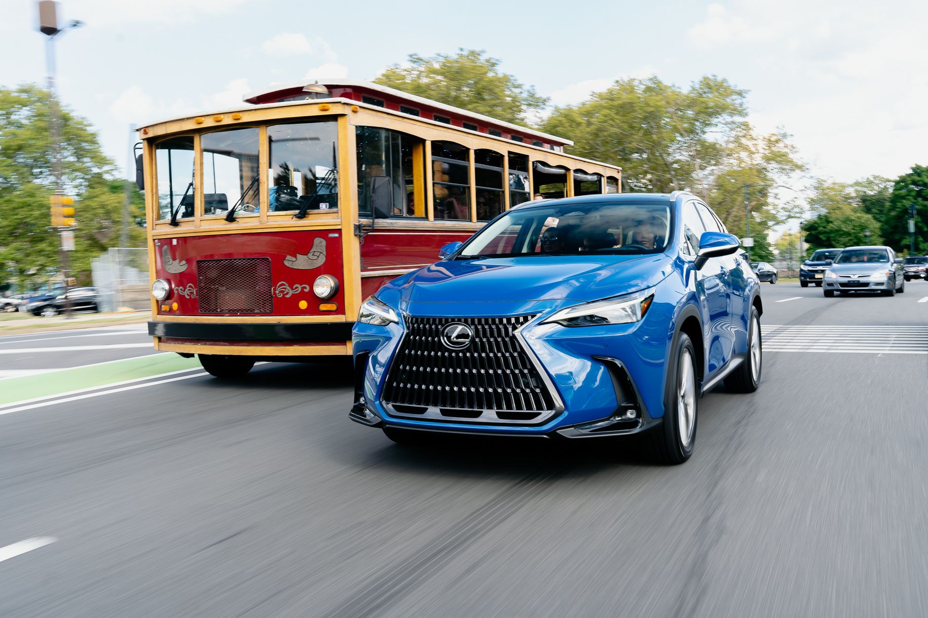 A blue Lexus driving alongside a vintage trolley in a bustling urban street setting.