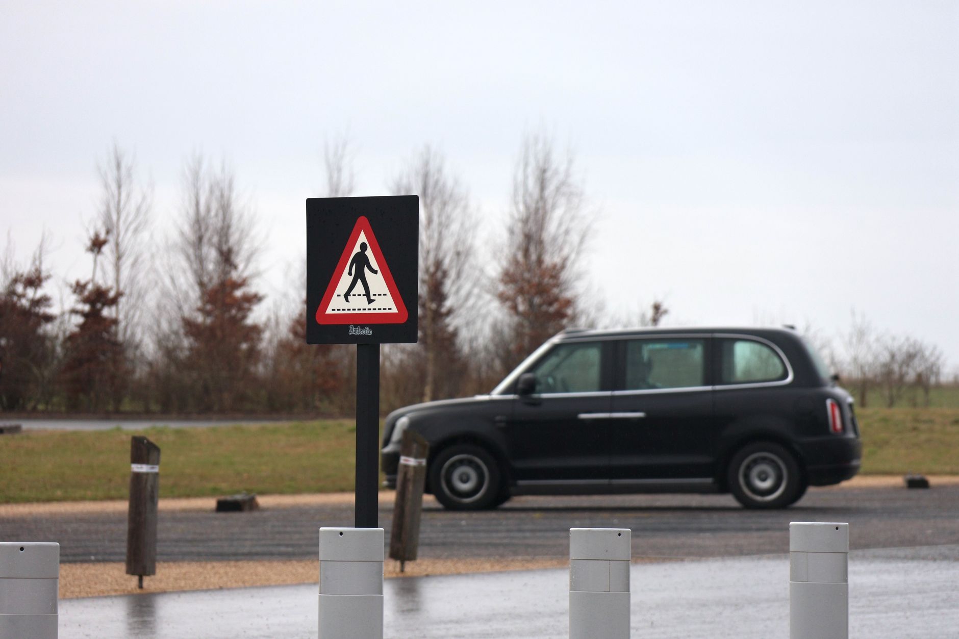Pedestrian crossing sign in foreground with black taxi parked on the road in a countryside setting.