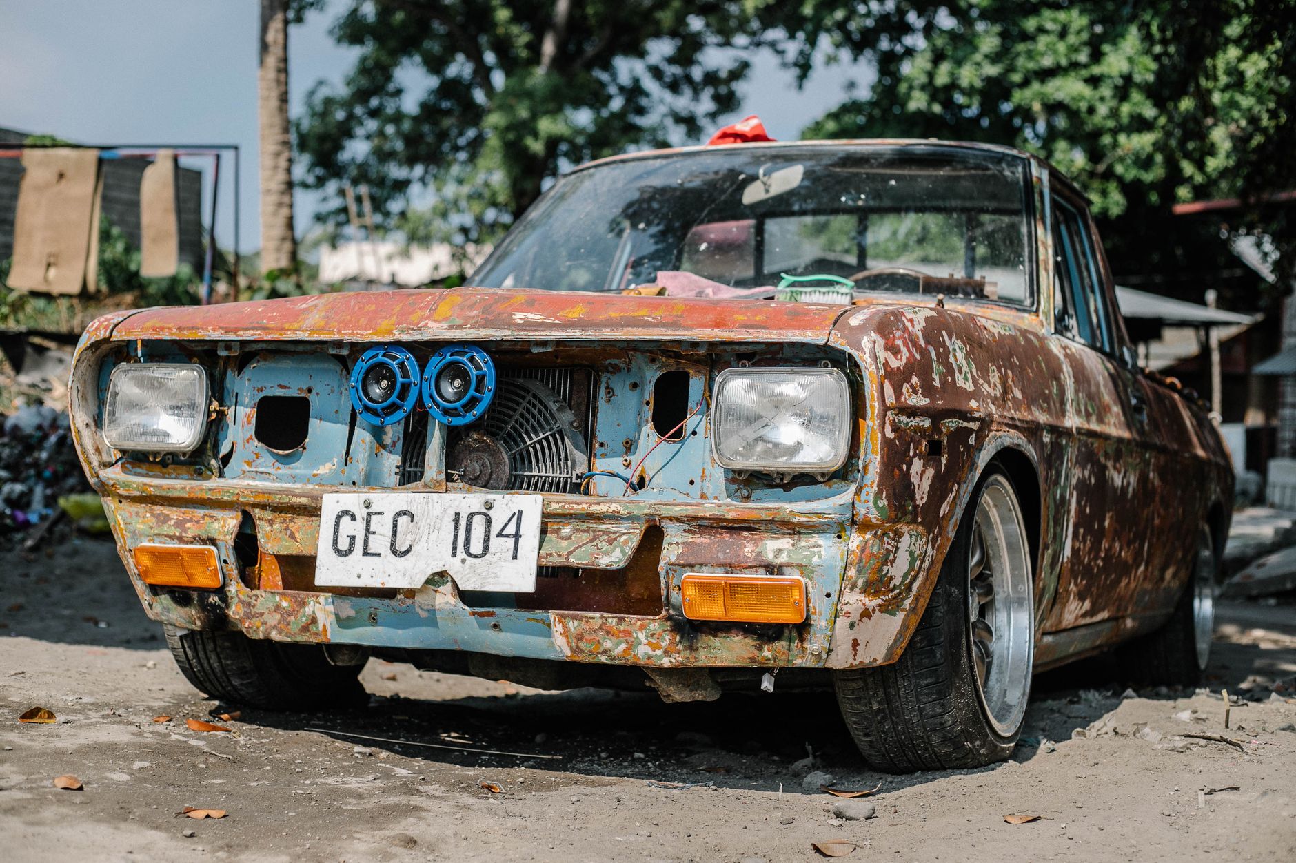 Aged and rusty vintage car parked outdoors on a sunny day, featuring peeling paint and visible wear.