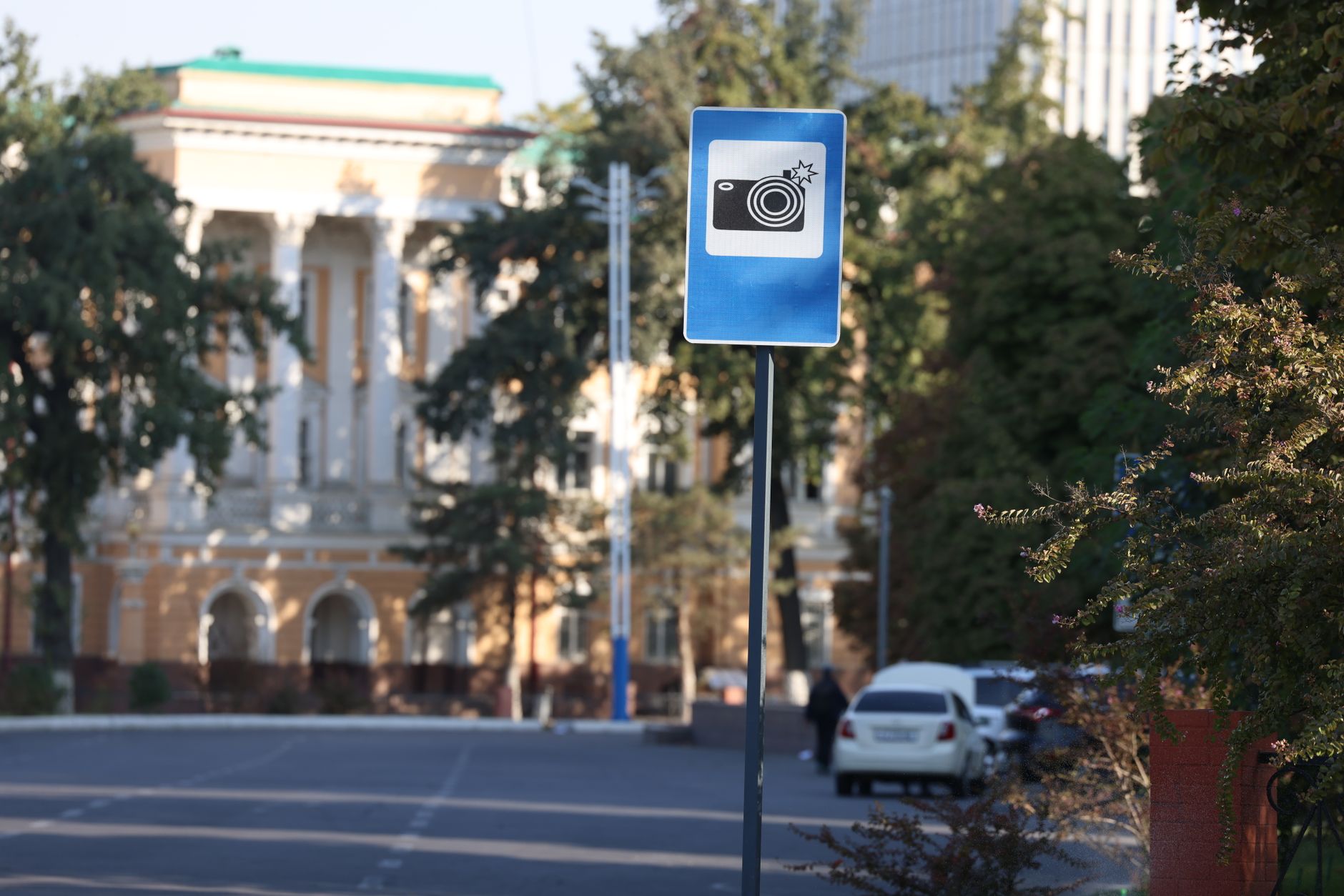 A blue traffic camera warning sign on a city street with blurred buildings in the background.