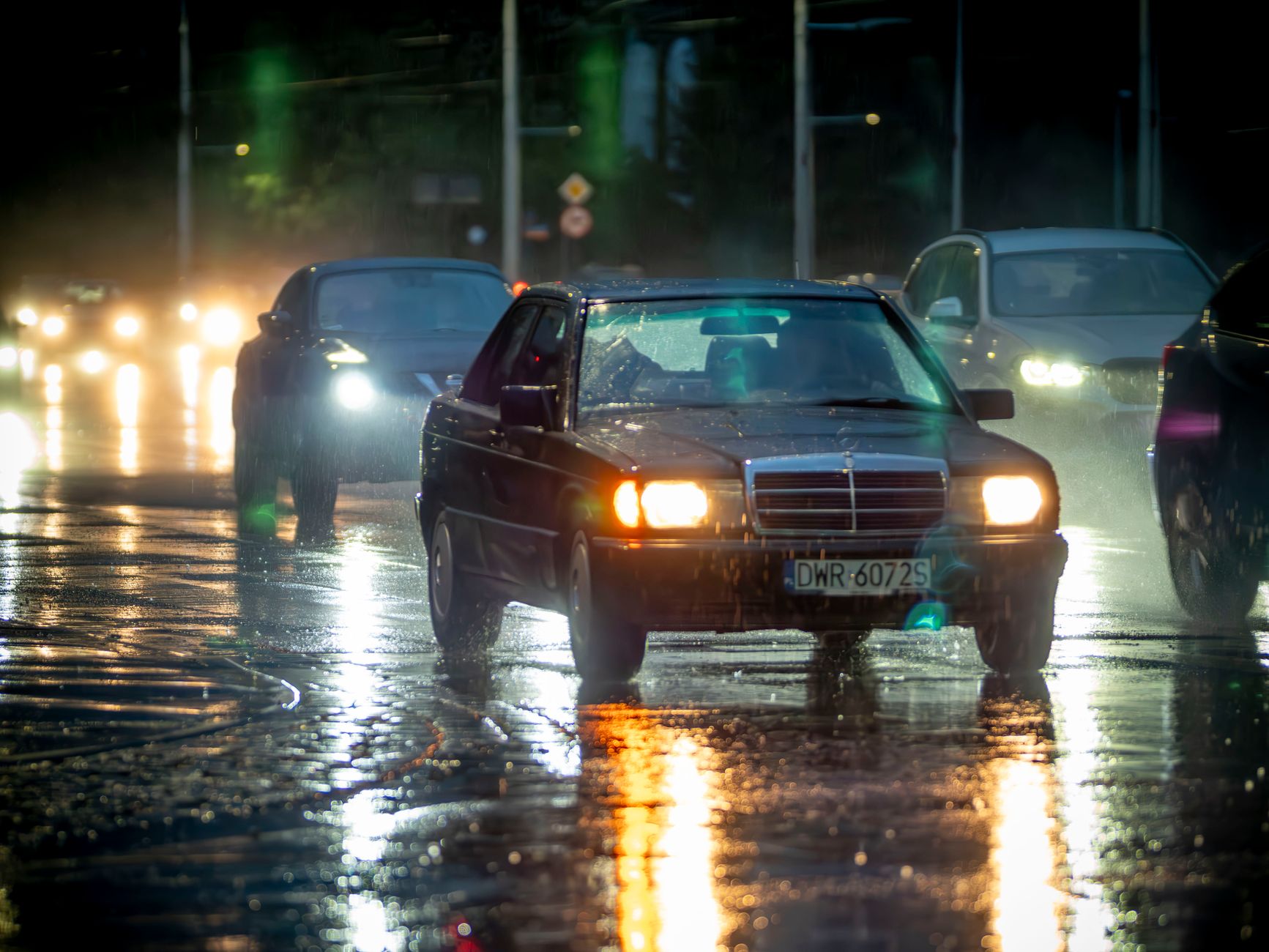 Cars drive through rainy night streets in Wrocław, Poland, reflecting vibrant city lights.