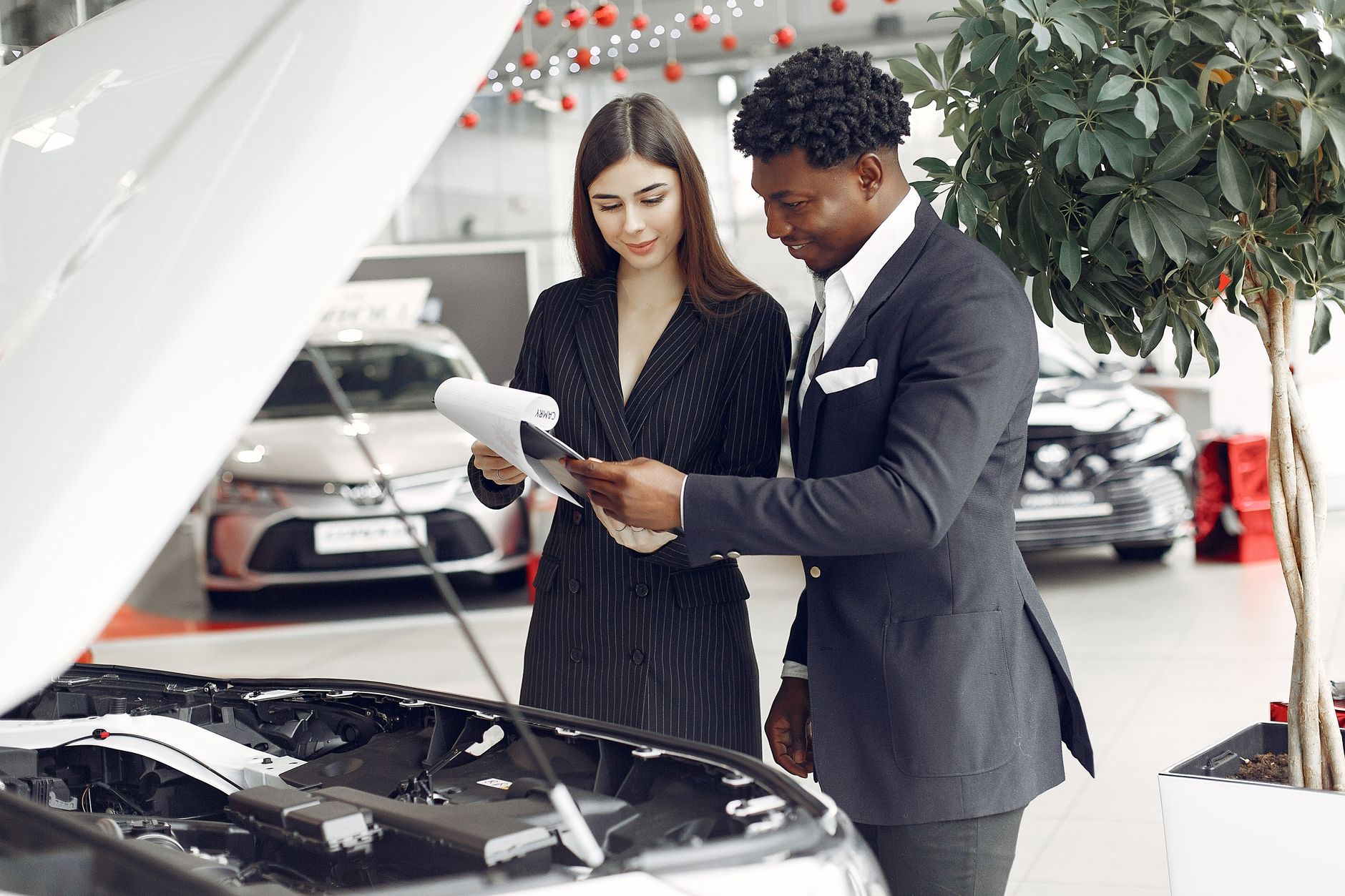 Confident smiling successful black businessman in classy formal suit standing near open motor hood in car showroom and discussing car charac