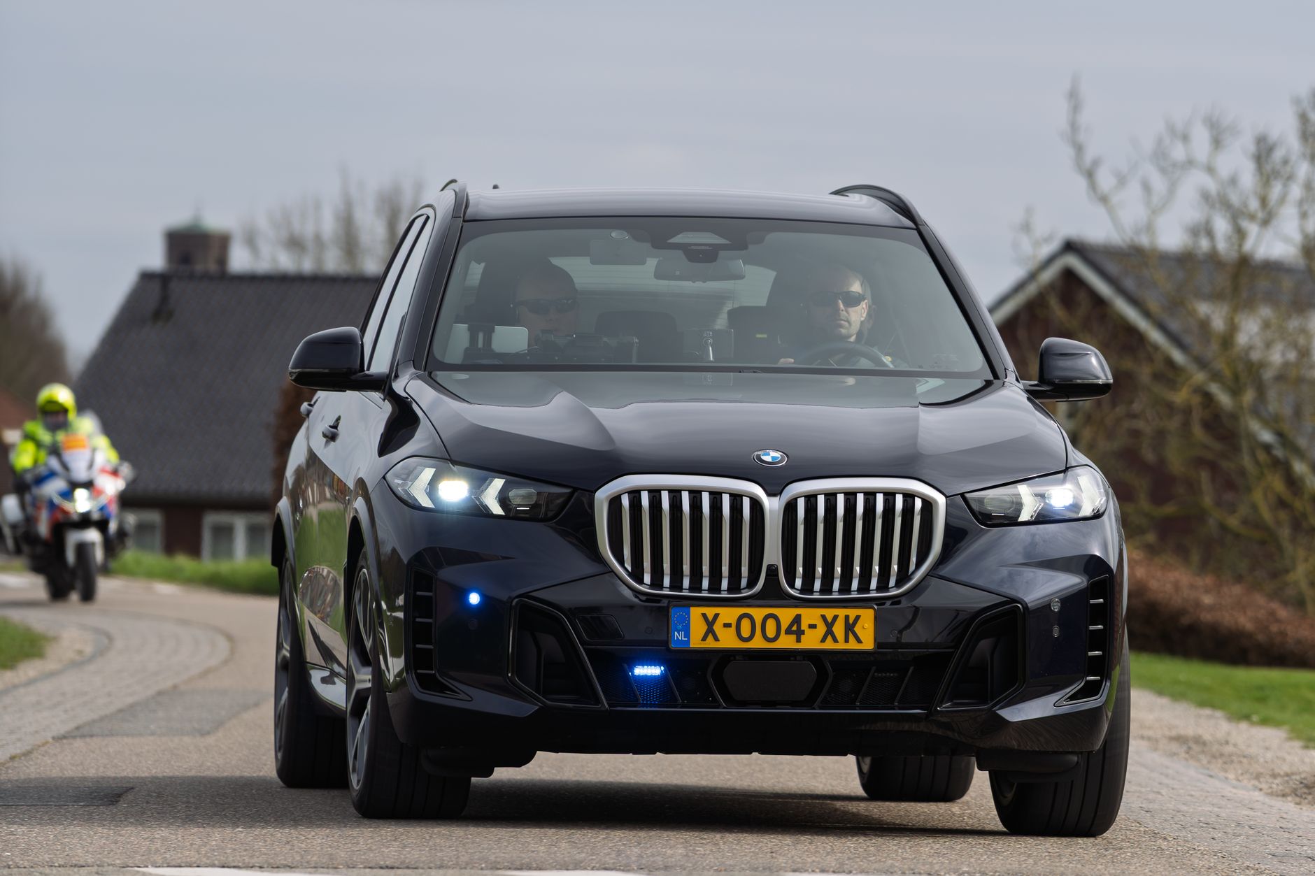 A sleek black luxury SUV travels on a rural road in the Netherlands with a motorbike escort.