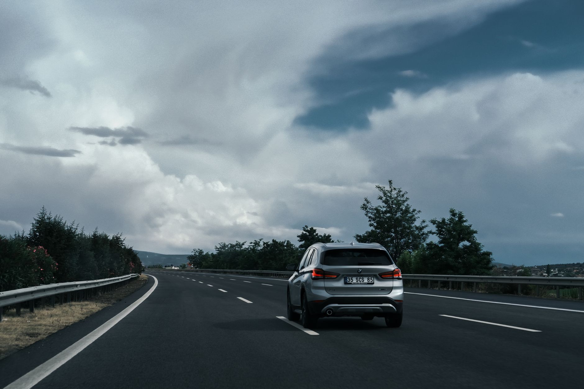 A silver SUV travels on a highway surrounded by lush greenery and dramatic clouds.