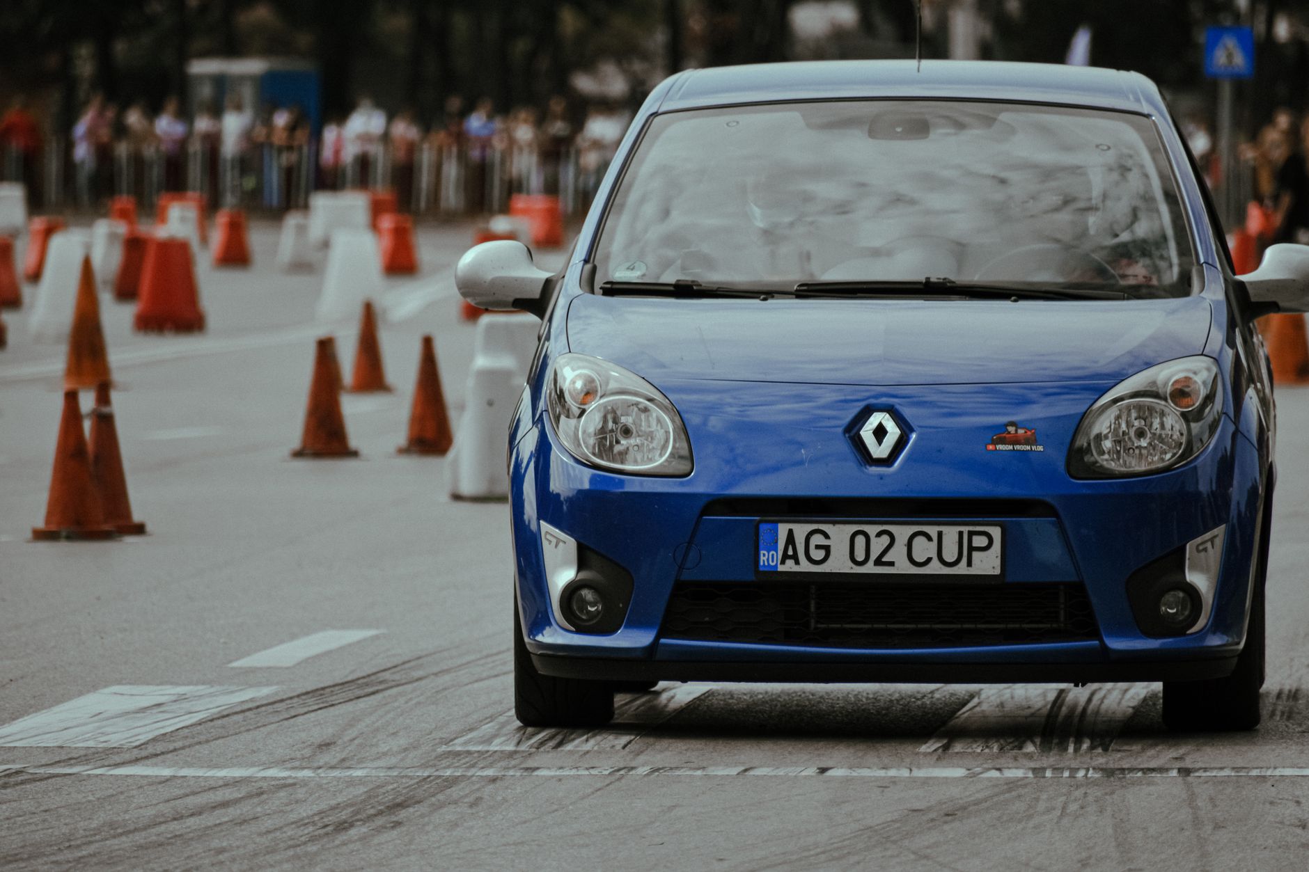 Modern blue automobile with headlights riding on pavement with marking lines near traffic cones on autodrome in city street with blurred bac