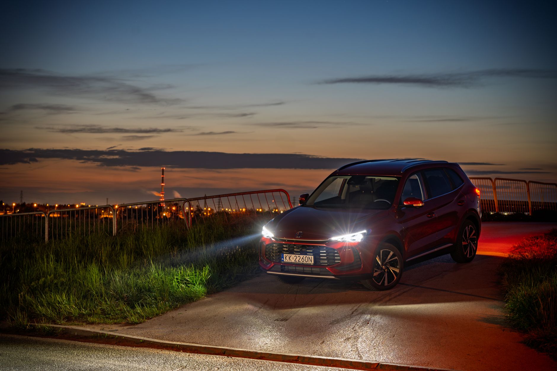 A sleek SUV with headlights on, parked at dusk near Kokotów, Poland, with a cityscape backdrop.