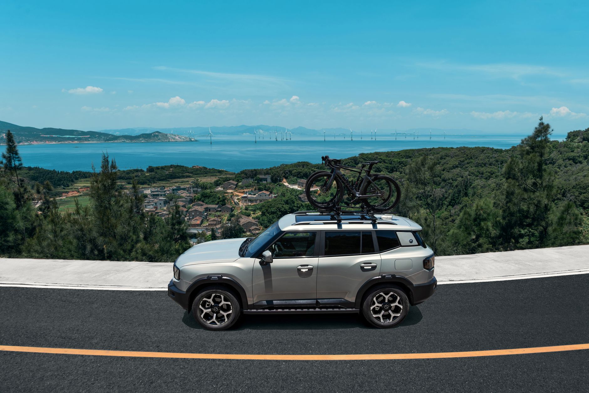 Silver SUV with bicycles on the roof rack driving along a coastal road surrounded by lush green hills and ocean view.