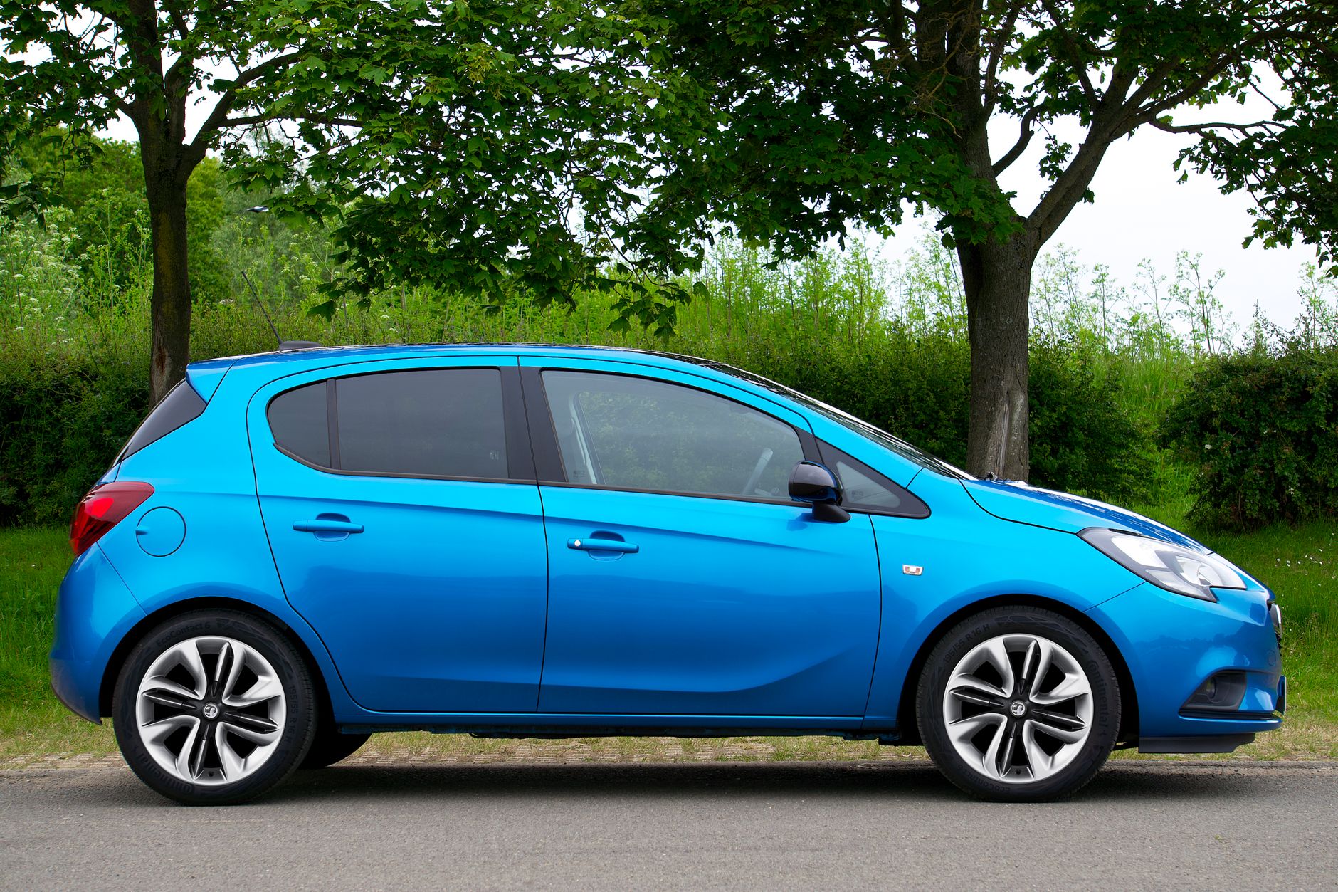 Side view of a bright blue compact car parked beside lush green trees and grass.