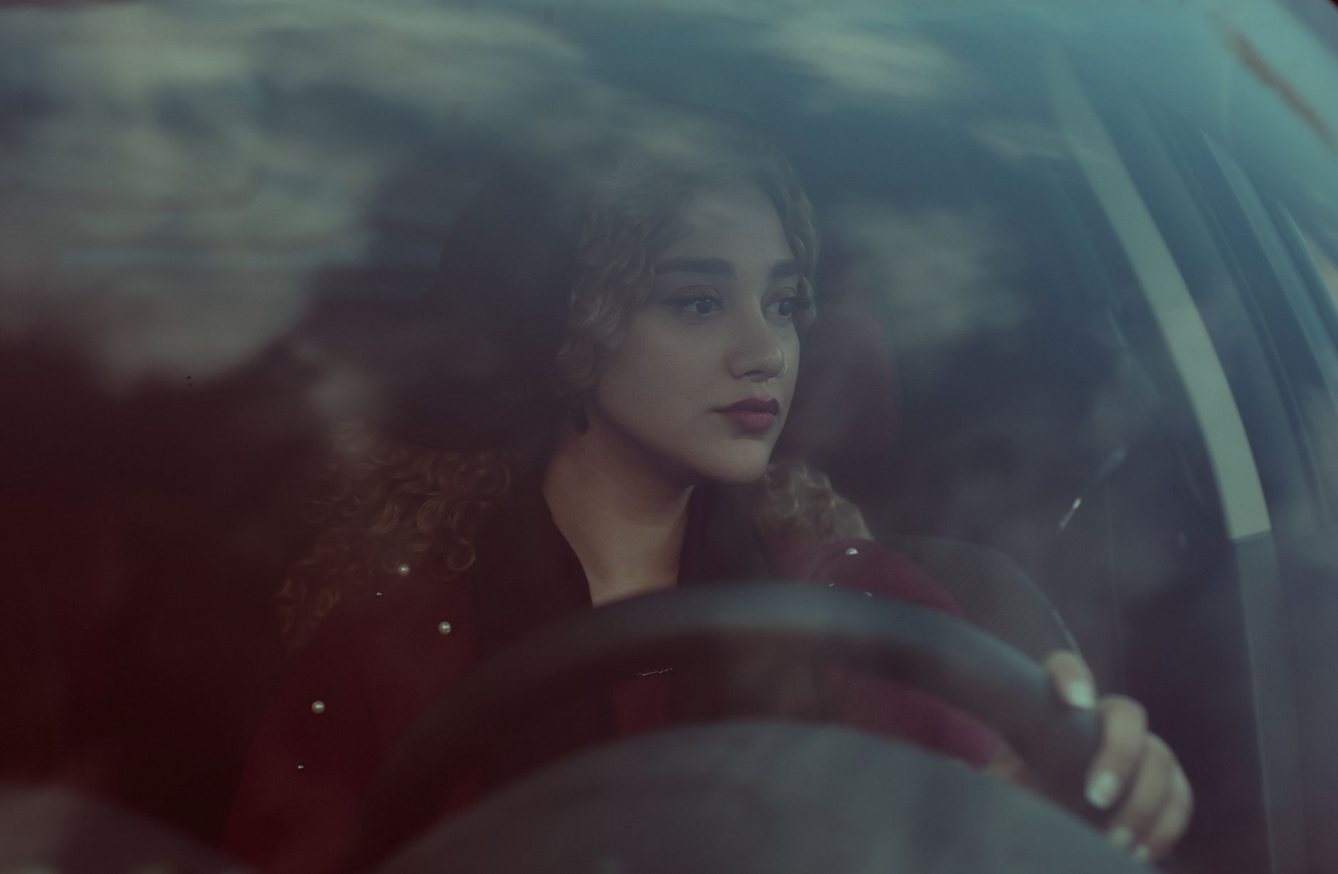 A young woman with curly hair focuses on driving through the city, reflecting her thoughtful expression.