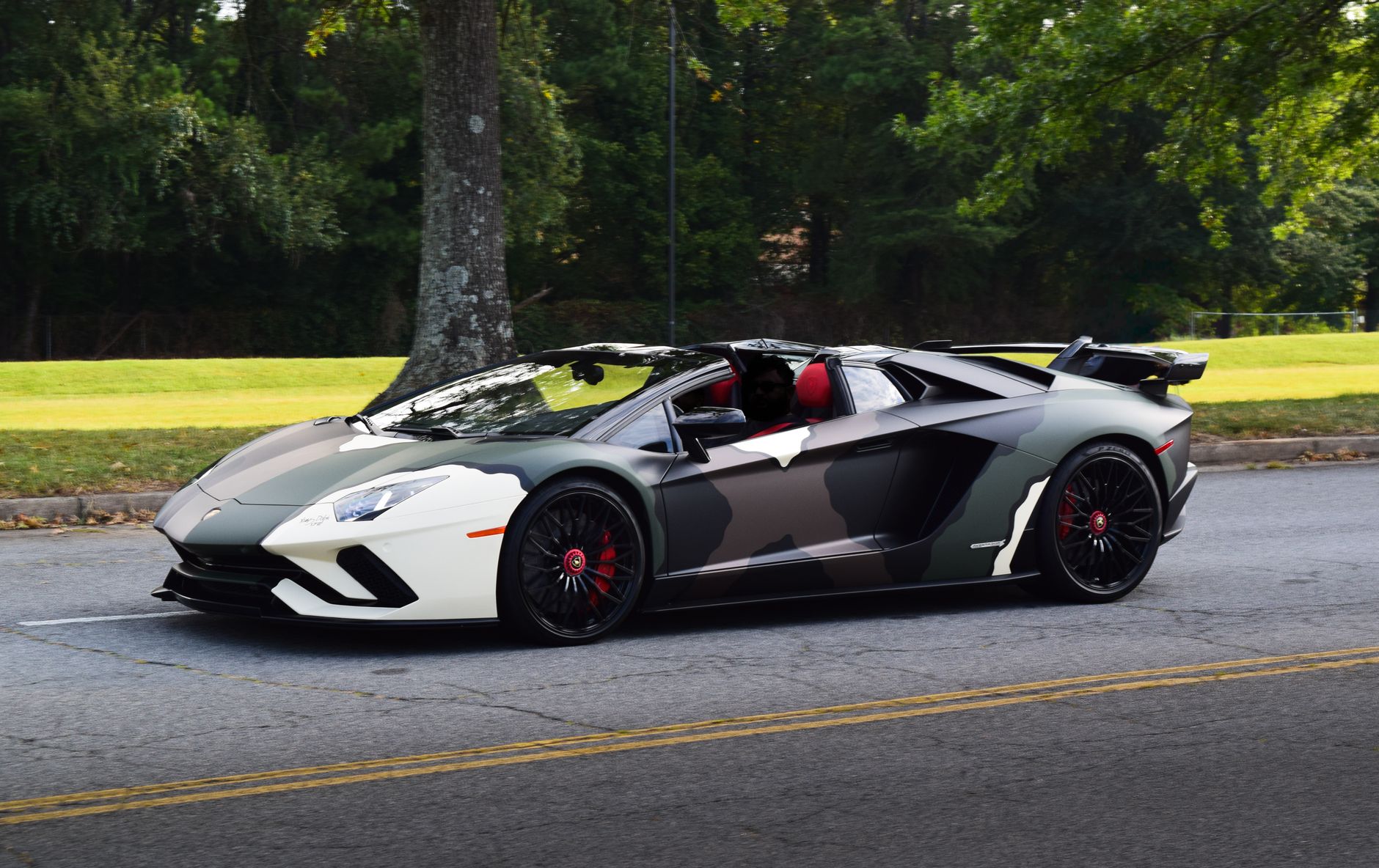 Camouflage-patterned luxury sports car cruising on a tree-lined street on a sunny day.
