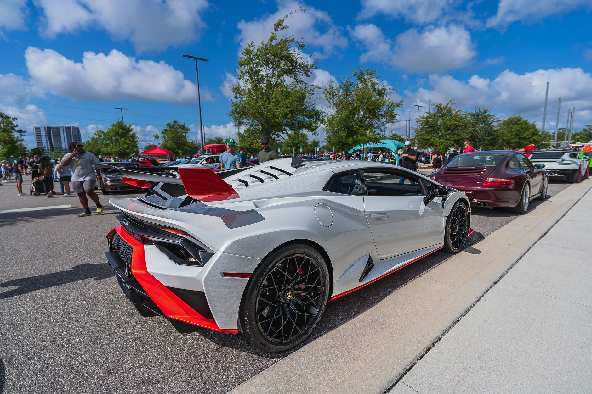 A lineup of high-performance sports cars at an outdoor car show on a sunny day, with a crowd gathered.